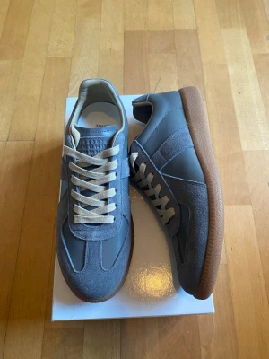 Grå Margiela Gats med mockadetaljer - (Helt Nya) Snygga grå sneakers med ovandel i både mocka och slätt läder, vita snören och klassisk gummisula i brunt. Skorna har en stilren och tidlös design med diskreta paneler och låg profil som passar till det mesta.