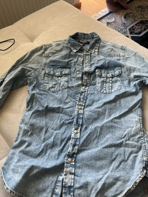 Ljusblå jeansskjorta med knappar - Säljer en ljusblå jeansskjorta med långa ärmar och tryckknappar framtill. Skjortan har två bröstfickor med lock och knapp, klassisk krage och en avslappnad passform. Perfekt för en cool och avslappnad stil.
