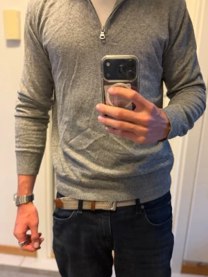Grå halvzip tröja Oscar Jacobson - Snygg grå stickad tröja från Oscar Jacobson med halv dragkedja framtill. Tröjan har ribbade muddar vid ärmslut och nederkant samt en klassisk passform. Perfekt till lager på lager och enkel att matcha med olika outfits.