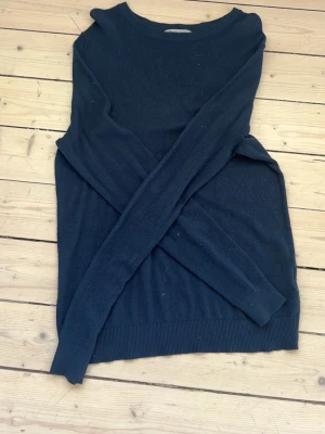 Mörkblå stickad tröja - En enkel och stilren mörkblå stickad tröja med rund halsringning och långa ärmar. Tröjan har ribbade muddar vid ärmslut och nederkant. Perfekt att matcha med jeans eller kjol för en avslappnad look.
