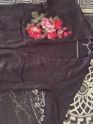 Svart kofta med broderade rosor - Mysig svart kofta med stora broderade rosor i rött, rosa och grönt på ena sidan. Koftan har två framfickor och är öppen framtill utan knappar. Perfekt för dig som gillar blommiga detaljer och vill ha något extra till din outfit.