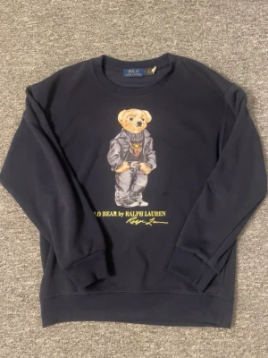Mörkblå Polo Bear sweatshirt - Mörkblå sweatshirt från Ralph Lauren med ikoniska Polo Bear-trycket framtill. Tröjan har rund halsringning, ribbade muddar och avslappnad passform. Perfekt för en chill och stilren look.