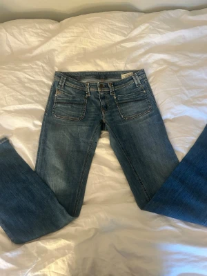 Disel jeans med låg midja - Säljer ett par klassiska blå bootcut jeans med låg midja och fem fickor. Jeansen har snygga slitningar och kontrastsömmar. Tillverkade i stretchigt denimtyg för skön passform. Perfekta till sneakers eller boots.