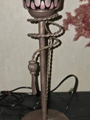 Dekorativ bordslampa  - Unik bordslampa med bronsfärgad metallfot och snirklig tvinnad detalj runt stommen. Lampan har en kupa i rosa och svart glas med dekorativa mönster. Perfekt som inredningsdetalj för dig som gillar vintage och annorlunda design.