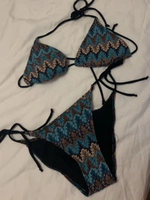 Mönstrad bikini från Design by Si - Säljer en snygg trekantsbikini från Design by Si med zigzag-mönster i blått, brunt, svart och vitt. Både överdel och underdel har knytband i sidorna och nacken för justerbar passform. Perfekt för strandhäng eller poolparty! Oanvänd, liten i storleken.