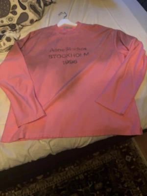 Rosa långärmad topp Acne Studios - Cool rosa långärmad topp från Acne Studios med tryck på bröstet där det står 'Acne Studios STOCKHOLM 1996'. Tillverkad i mjuk bomull med rund halsringning och relaxed fit. Perfekt för dig som gillar streetwear och skandinavisk design.                                                    Ärmlängd (axelsöm till ärmslut) 63–66 cm.         Längd från axel (vid halsen) till nederkant (axel till midja)69–73 cm