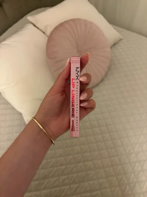 NYX Lip Lingerie läppstift nude - NYX Lip Lingerie är ett flytande läppstift i nude-rosa ton. Förpackningen är smal och rosa med tydlig logga. Ger en matt finish och är enkel att applicera tack vare den smidiga applikatorn. Perfekt för dig som gillar naturliga läppar med en hint av färg.