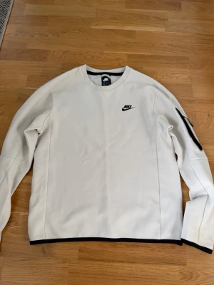 Vit Nike sweatshirt med svart logga - Snygg vit sweatshirt från Nike med svart logga på bröstet och svarta detaljer vid ärmslut och nederkant. Tröjan har rund halsringning och en diskret ficka med dragkedja på ena ärmen. Perfekt för en clean och sportig look.