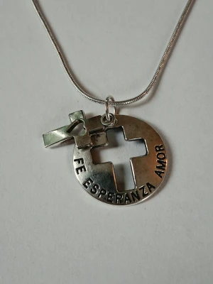Silverfärgat halsband med kors och text - Snyggt halsband i silverfärgad metall med ett runt hänge där ett kors och ett hjärta är utskurna. Runt hänget står texten 'FE ESPERANZA AMOR' som betyder tro, hopp och kärlek. Kedjan är tunn och smidig. Materialet rostfritt stål. 