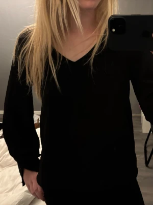 Svart blus med v-ringning - Stilren svart blus med v-ringad hals och långa ärmar. Ärmsluten har dekorativa knappar som ger en extra detalj till looken. Perfekt att matcha med både jeans och kjol. Materialet ser ut att vara lätt och något genomskinligt.