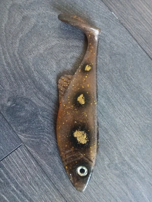 Mjukfiskbete i brunt med glitter - Säljer ett mjukfiskbete i brun plast med svarta och guldglittriga detaljer. Betet har realistisk fiskform med fenor och ett stort öga, perfekt för fiske efter rovfisk. Passar till olika typer av fiske och är lätt att fästa på kroken. Hemmagjord 