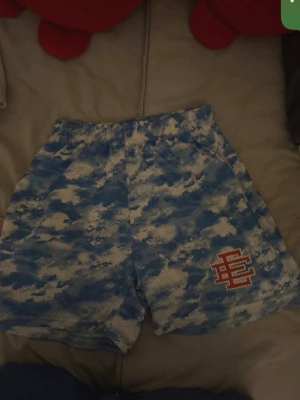 Blå camo meshshorts med tryck - Säljer ett par blå och vita meshshorts med camouflagemönster och elastisk midja. Shortsen har ett rött tryck på ena benet och är perfekta för träning eller chill. Lätta och luftiga med normal passform.