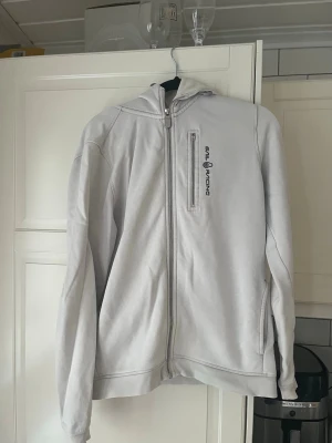 Vit hoodie från Sail Racing - Ljusgrå hoodie från Sail Racing med dragkedja framtill och en bröstficka med logga. Tillverkad i mjukt material med borstad insida för extra komfort. Klassisk huva och ribbade muddar vid ärmslut och nederkant. Perfekt för en avslappnad stil.