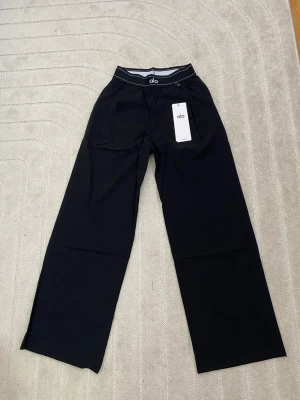 Svarta Suit Up mjukisbyxor från Alo S - Snygga svarta mjukisbyxor från Alo med breda ben och elastisk midja. Byxorna har fickor framtill och en diskret logga på benet. Perfekta för en chill och avslappnad stil. Materialet känns mjukt och bekvämt mot huden.