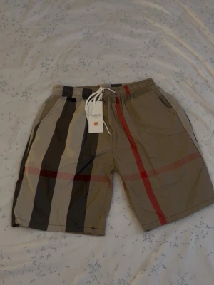 Burberry shorts  - Snygga shorts i beige med breda ränder i svart, brun och röd. Shortsen har elastisk midja med vit snörning och sidofickor. Perfekta för sommaren och ger en trendig look med sitt klassiska mönster.