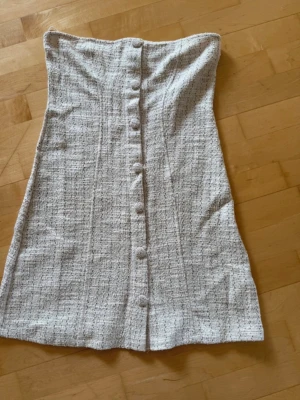 Beige tweedklänning  - Säljer en snygg beige tweedklänning från Gina Tricot. Klänningen är axelbandslös, har knappar framtill och är i en kort modell. NY med prislapp!