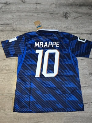 Frankrike Mbappé matchtröja blå - Säljer en blå matchtröja från Nike med Frankrikes landslagsemblem och nummer 10 Mbappé på ryggen. Tröjan har korta ärmar, vit krage och snyggt mönster i olika blå nyanser. Perfekt för dig som älskar fotboll och vill stötta Frankrike.