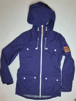 CLWR Colour Wear skid / snowboardjacka / outdoor - Snygg skid-/snowboardjacka från CLWR / Colour Wear i blå/lila färg med vita detaljer. Huva, hög krage, flera fram- och innerfickor, snölås/snökjol, dragsko samt ventilation med dragkedja under armarna. Alla dragkedjor, knappar och snörspännen fungerar. Fin snow/outdoor-stil som även passar som vardagsjacka/streetwear.  Storlek: M Mått: bröst 48 cm, längd 80 cm, ärm 53 cm. Begagnat skick enligt bilder. Väldigt lite använd. 