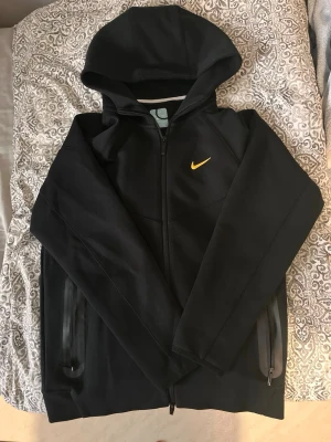 Svart Nike nocta tech fleece (size M men passar som S) - Snygg svart hoodie från Nike med hel dragkedja och gul logga på bröstet. Utrustad med huva och två praktiska fickor med dragkedja. Mjukt och sportigt material som är perfekt för dig som gillar sportig stil och chill stil.