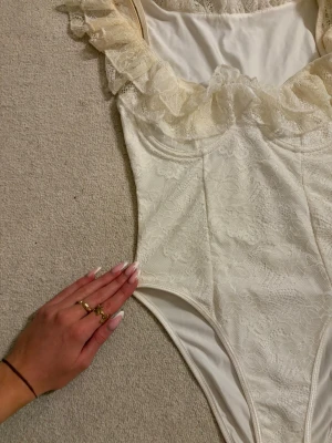 Vit spetsbody med volang - Snygg vit body i spets med romantiska volangdetaljer runt urringningen. Bodyn har smala axelband och är tillverkad i ett stretchigt spetsmaterial som ger en feminin look. Perfekt för dig som gillar detaljer och en elegant stil.