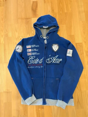 Blå hoodie med broderade detaljer - Snygg blå hoodie med dragkedja och grå muddar. Flera broderade patches och text på både fram- och baksida, bland annat 'Cote d'Azur' och en stor siffra 5 med 'Monte-Carlo' på ryggen. Utrustad med huva och fickor framtill. Perfekt för en sportig och avslappnad stil.
