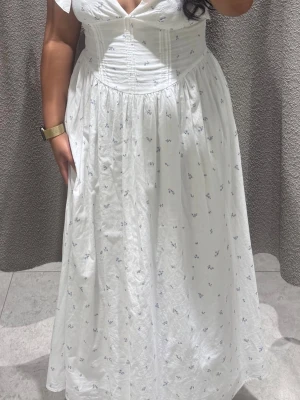 Gina tricot Vit långklänning  - Söt vit långklänning med små blå blommor och markerad midja. Klänningen har korta ärmar, v-ringning och en lätt rynkad kjol som ger en romantisk känsla. Perfekt för varma dagar och somriga tillfällen.