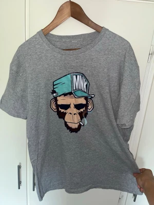 Grå t-shirt med apa och keps - Säljer en grå t-shirt med ett stort tryck av en apa med blå keps och cigarett på bröstet. T-shirten har rund hals och korta ärmar. Perfekt för dig som gillar streetwear och vill sticka ut lite extra.