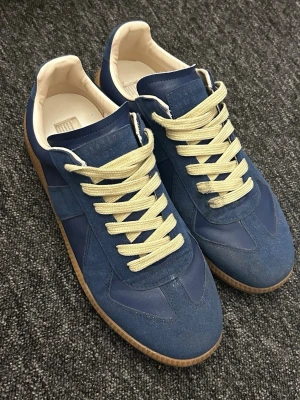 Blå sneakers med beige snören - Snygga blå sneakers i mocka och läder med beige snören och vit insida. Skorna har en klassisk låg siluett och gummisula som ger en retrovibe. Perfekta för dig som gillar stilrena och tidlösa sneakers.