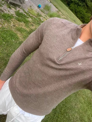 Morris merino ull quarter zip  - Snygg beige stickad tröja med halv dragkedja och liten vit broderad detalj på bröstet. Tröjan har lång ärm och ribbade muddar vid ärmslut och nederkant. Perfekt för lager-på-lager och enkel att matcha med olika stilar.