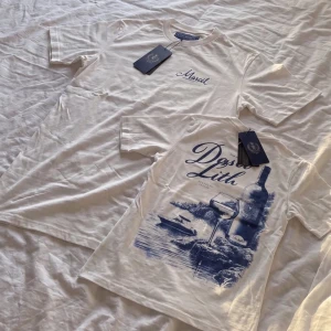 Vit t-shirt med blått tryck - Säljer en vit t-shirt med blått tryck på ryggen som föreställer vinflaska, glas och landskap. Framtill står det 'Mariel' broderat i blått. Klassisk passform och rund hals. Perfekt för dig som gillar stilrena plagg med unik detalj.