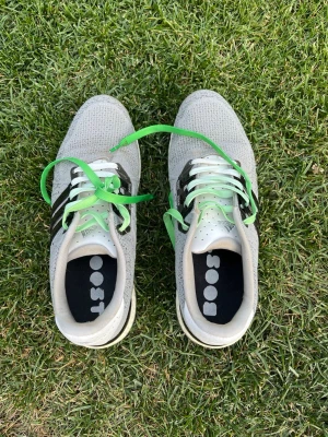 Grå sneakers med gröna snören - Snygga grå sneakers med stickad ovandel och svarta detaljer på sidorna. Skorna har gröna och vita skosnören samt en vit sula. Perfekta för dig som gillar en sportig och modern stil. Insidan har svart innersula med text.