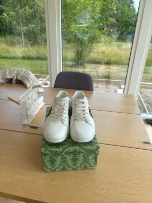 Vita sneakers med färgade detaljer - Snygga vita sneakers i läder med klassisk låg profil. Skorna har unika detaljer i rött och grönt ormskinnsmönster på hälen, vilket ger en cool kontrast. Perfekta för dig som vill sticka ut med stilrena men ändå speciella skor.