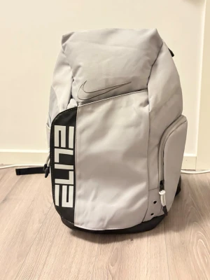 Nike Elite grå ryggsäck - Snygg grå Nike Elite ryggsäck med svart och vit detalj på framsidan. Ryggsäcken har flera fack, vadderade axelband och dragkedjor. Tillverkad i slitstarkt material och har tydlig Nike-logga både fram och på axelbanden. Perfekt för skola, träning eller gym.