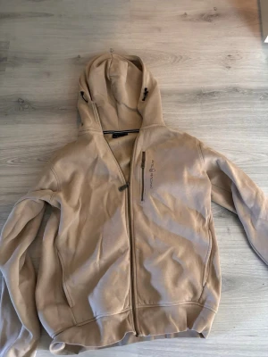 Beige hoodie med dragkedja - Säljer en beige hoodie med dragkedja framtill och huva med dragsko. Hoodien har en bröstficka med dragkedja och diskret broderad text på bröstet. Tillverkad i mjukt material, perfekt för chill dagar.
