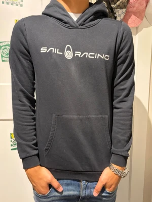 Svart  hoodie från Sail Racing - Mörkblå hoodie från Sail Racing med vit logga tryckt på bröstet. Klassisk känguruficka framtill och huva. Tillverkad i mjukt material och har ribbade muddar vid ärmslut och nederkant. Perfekt för en avslappnad och sportig stil.