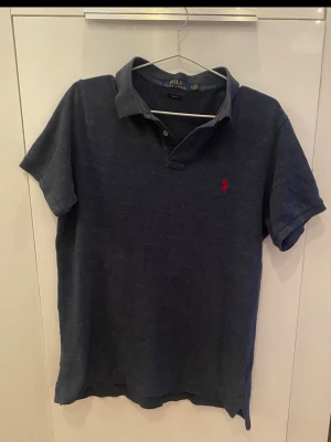 Marinblå piké från Polo Ralph Lauren - Klassisk marinblå pikétröja från Polo Ralph Lauren med röd broderad logga på bröstet. Slim fit-modell med korta ärmar och krage. Tillverkad i mjuk bomull som känns skön mot huden. Perfekt till jeans eller shorts för en clean och stilren look. Den är L men har krymt till en M 