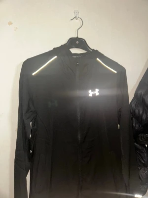 Under Armour windbreaker - Svart träningsjacka från Under Armour med hel dragkedja, huva och reflekterande detaljer på axlarna. Jackan har två sidofickor och är tillverkad i ett lätt och funktionellt material som passar perfekt för löpning eller gym.