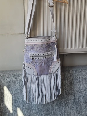 Vit Axelväska crossbody med fransar och nitar - Säljer en unik vit axelväska med långa fransar, silverfärgade nitar och spetsdetaljer. Väskan har justerbar axelrem, dragkedja upptill och en mindre ficka framtill med dragkedja. Perfekt för dig som gillar boho-stil och vill sticka ut.