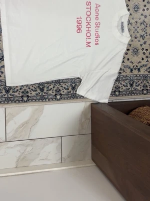 Vit Acne Studios t-shirt med tryck - Vit t-shirt från Acne Studios med rosa tryck framtill där det står 'Acne Studios STOCKHOLM 1996'. Klassisk passform och rund halsringning. Perfekt för en clean och stilren look.Tvättlappen är avklippt