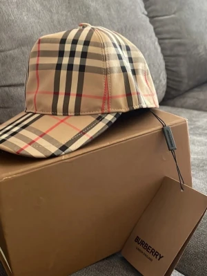 Burberry beige klassisk rutig keps - Säljer en snygg keps från Burberry i deras ikoniska beigea rutiga mönster med svarta, vita och röda detaljer. Justerbar rem bak med tryckknappar och Burberry London England-märke. Perfekt accessoar för dig som gillar streetstyle och lyxig känsla. Ingen box medföljer