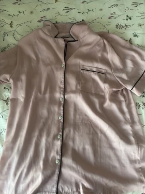  Pyjamas skjorta från Anna Field  - Snygg ljusrosa pyjamasskjorta med korta ärmar och svarta kontrastkanter. Skjortan har knäppning framtill och en bröstficka med svart kant. Tillverkad i ett mjukt, glansigt material som känns lyxigt mot huden. Perfekt för mysiga hemmakvällar.
