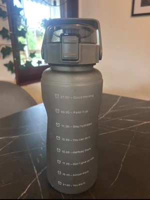 Grå vattenflaska med tidsmarkeringar - Säljer en grå vattenflaska i plast med motiverande tidsmarkeringar och citat längs sidan. Flaskan har ett praktiskt lock med bärhandtag och säkerhetslås, perfekt för att hålla koll på vätskeintaget under dagen.