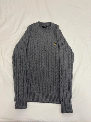 Tröja från Lyle & Scott - Säljer en grå kabelstickad tröja med rund hals och ribbade muddar. Tröjan har ett gult broderat märke på bröstet och är långärmad. Perfekt för kyliga dagar och enkel att matcha med jeans eller kjol.