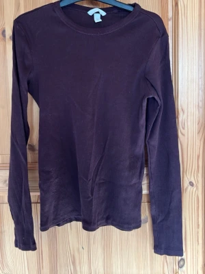 Mörkbrun ribbad långärmad topp - En enkel och stilren mörkbrun långärmad topp med rund halsringning. Toppen är ribbad och har en slim passform som sitter snyggt på kroppen. Perfekt att matcha med jeans eller kjol för en clean look.