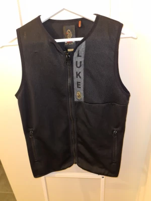 Svart väst från Luke Sport - Snygg svart väst från Luke Sport med dragkedja framtill och två fickor med dragkedja. Västen har en diskret logga och texten LUKE på bröstet. Perfekt för lager på lager och sportig stil. Nypris 1300
