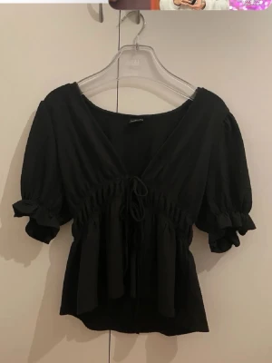 Svart blus med snörning Gina Tricot - Supersöt svart blus från Gina Tricot med korta puffärmar, v-ringning och snörning i midjan. Blusen har volangdetaljer vid ärmslut och en lätt rynkad struktur som ger en feminin look.