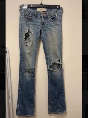 Hollister jeans - Säljer ett par ljusblå bootcut jeans med stora slitningar och hål på båda benen. Jeansen har klassisk femficksdesign, låg midja och snygga broderade detaljer på bakfickorna. Perfekta för en avslappnad och trendig streetstyle-look.