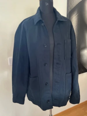 Mörkblå overshirt från John Henric  - Stilren mörkblå overshirt med klassisk krage och knäppning framtill. Tre stora utanpåfickor ger en avslappnad look. Tillverkad i ett kraftigare tyg som passar perfekt som lager. Enkel och tidlös design som funkar till det mesta.