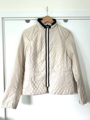 Soyaconcept - beige quiltad jacka med svarta detaljer storlek M - Stilren beige / cremefärgad/ vit lätt vadderad jacka från Soyaconcept i storlek M. Smickrande modell med quiltat mönster på kroppen och perforerade ärmar. Jackan har en ståkrage och stängs med dragkedja framtill. Dekorativa dragkedjor i ärmsluten. Diskreta fickor framtill som stängs med dragkedja. Snygg detalj i kontrastfärg (svart) runt dragkedja, krage och vid ärmsluten. Perfekt allvädersjacka. Mycket gott skick, som ny!