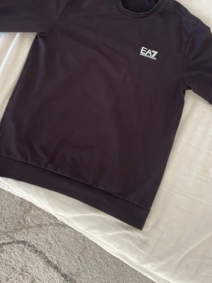 Svart sweatshirt från EA7 - Snygg svart sweatshirt från EA7 med rund halsringning och diskret logga på bröstet. Tröjan har en klassisk passform och ribbade muddar vid ärmslut och nederkant. Perfekt för en clean och stilren look.